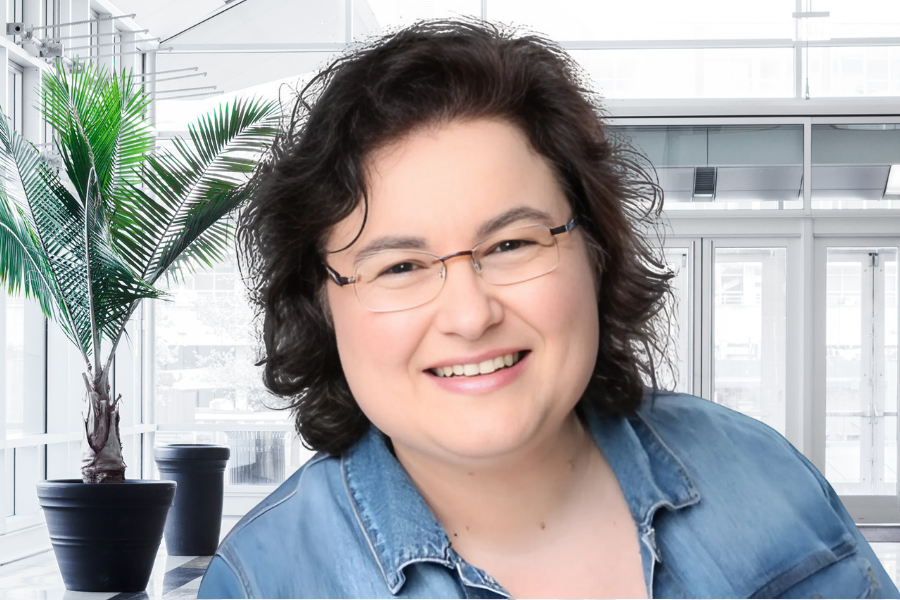 Simone Winkler lächelt freundlich in die Kamera. Sie trägt eine Brille und ein Jeanshemd. Im Hintergrund ist eine helle, moderne Büroumgebung mit großen Fenstern und Palmenpflanzen zu sehen.