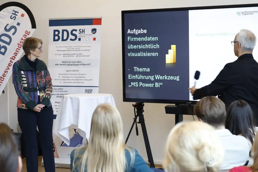 Vortragssituation bei einer BDS-Schleswig-Holstein-Veranstaltung: Referent präsentiert auf einem Bildschirm Inhalte zur Datenvisualisierung mit MS Power BI, Teilnehmende hören zu. BDS-SH-Roll-up im Hintergrund.